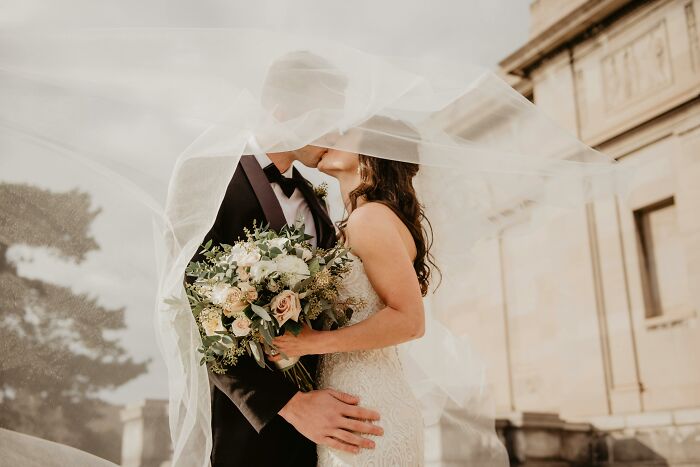 Pareja besándose bajo el velo en una boda, con el novio sosteniendo un ramo.