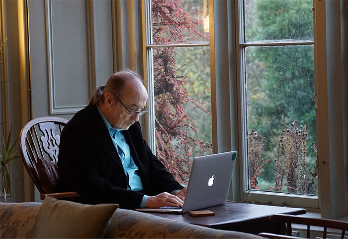 Hombre mayor escribiendo en computadora portátil junto a la ventana, reflejando problemas al publicar en redes sociales.