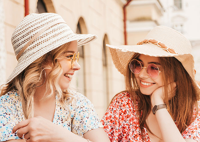 Dos amigas sonriendo, usando sombreros veraniegos y gafas de sol, hablando alegremente en una calle soleada.