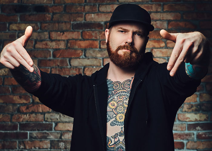 Hombre con tatuajes y gorra negra, gesticulando frente a un muro de ladrillos, símbolo de amistad y odio.