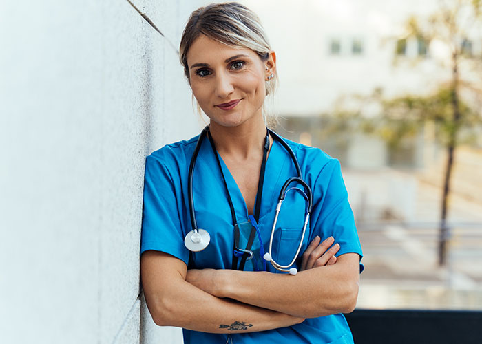 Mujer en uniforme médico azul junto a una pared, con fonendoscopio al cuello.