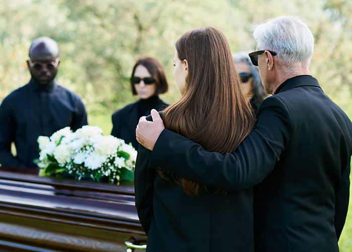 Personas en un funeral, vestidos de negro, rodeando un ataúd con flores blancas.