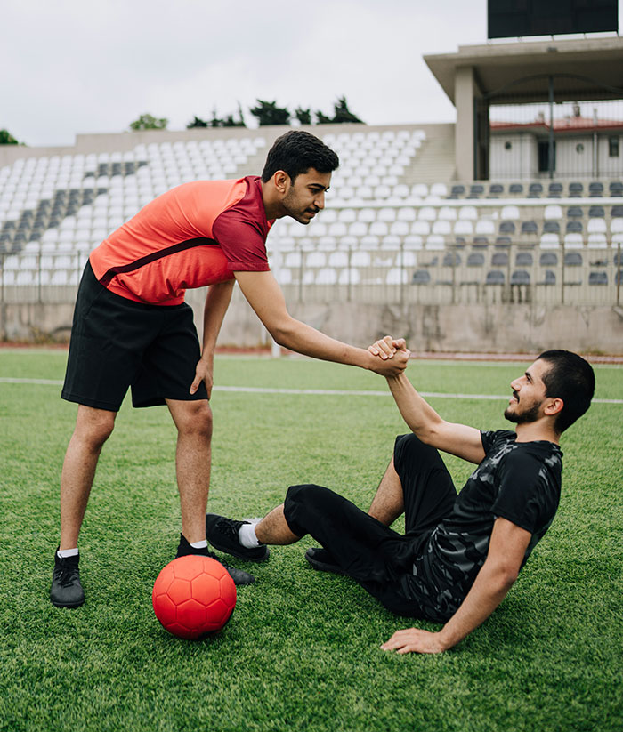 Jóvenes jugando al fútbol en un campo, uno ayudando a otro a levantarse, mostrando compañerismo.
