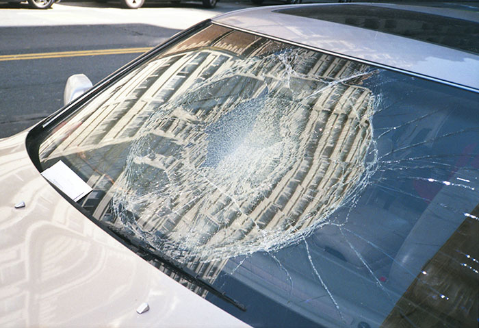 Parabrisas de auto con vidrio roto por impacto, reflejando edificios, relacionado con personas que siguen vivas por decisión espontánea.