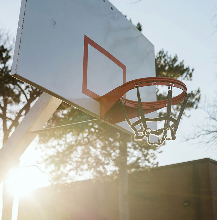 Aro de baloncesto al aire libre con luz solar y árboles de fondo, imagen relacionada con personas que aún siguen vivas.
