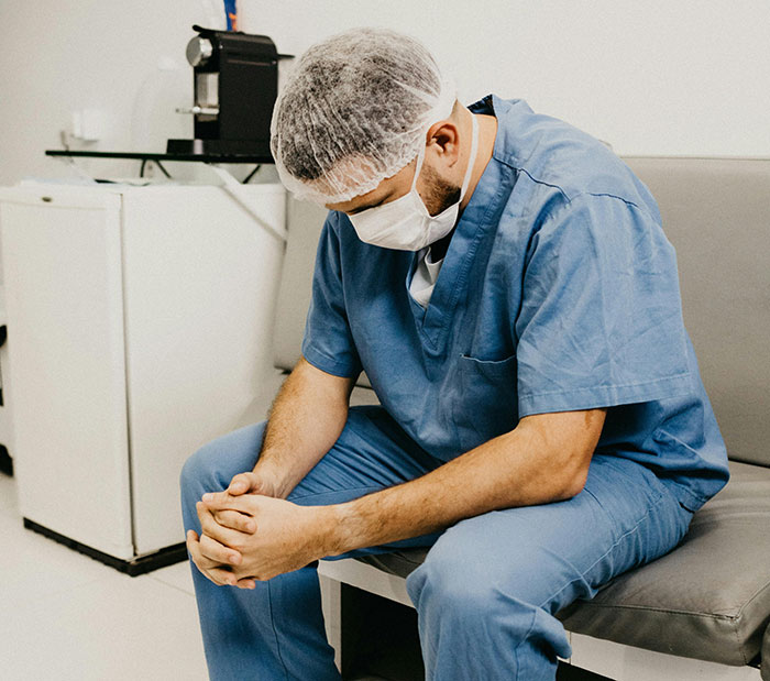 Médico sentado con uniforme azul y gorro quirúrgico, reflexionando sobre el cuerpo humano en una sala de espera.
