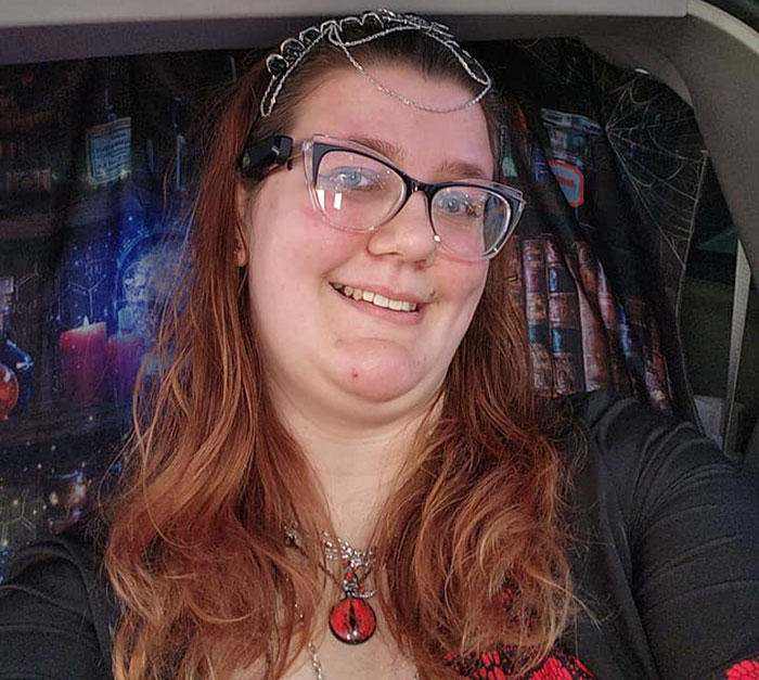 Smiling tall woman with glasses and jewelry, seated, colorful backdrop.