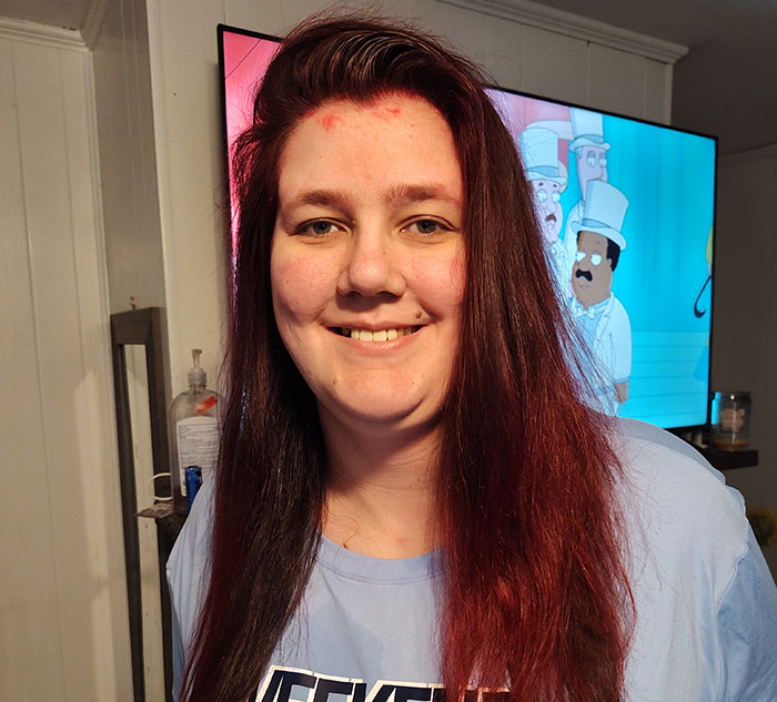 Smiling tall woman with long hair, standing indoors with a cartoon background on the TV.