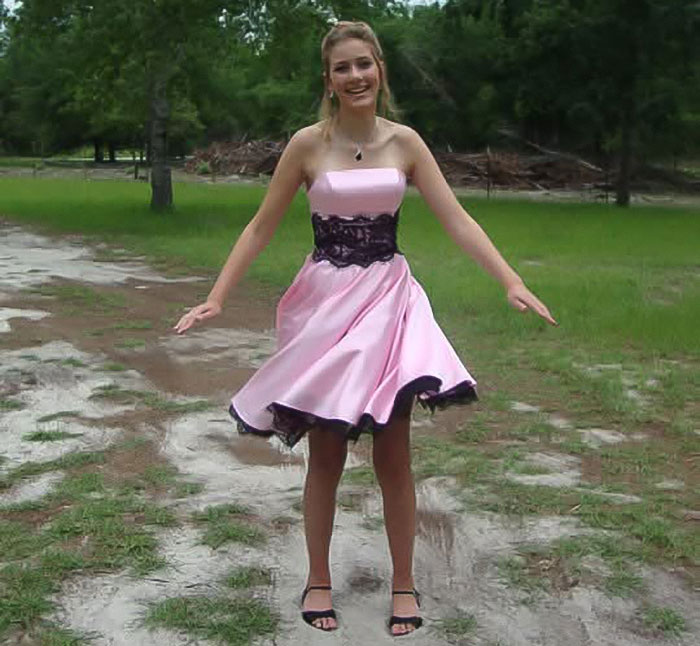 Tall cisgender woman in a pink dress standing outdoors on a dirt path with grass and trees.