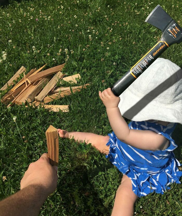 Bebé en el césped sosteniendo un hacha juguete mientras un adulto ofrece un trozo de madera, escena divertida.