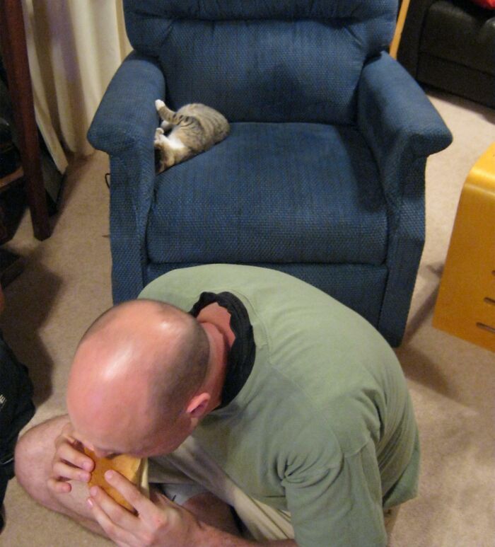 Un marido divertido haciendo una broma al frente de un sillón azul con un gato dormido.