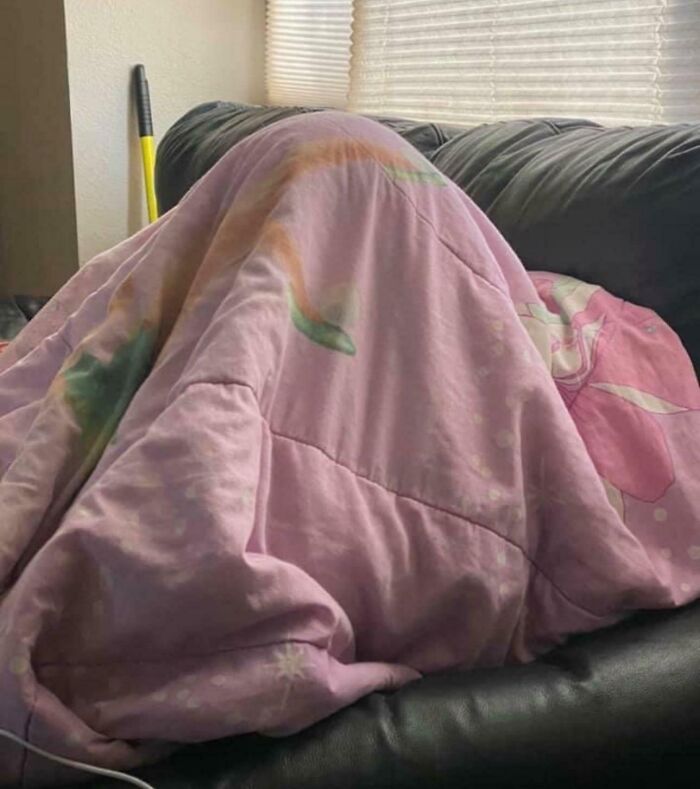 Novio divertido escondido bajo una manta morada en el sofá, haciendo reír a su pareja.