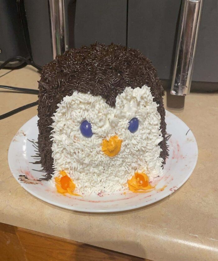 Pastel de repostería decorado como un pingüino con glaseado blanco, negro y detalles naranja y azul en un plato blanco.