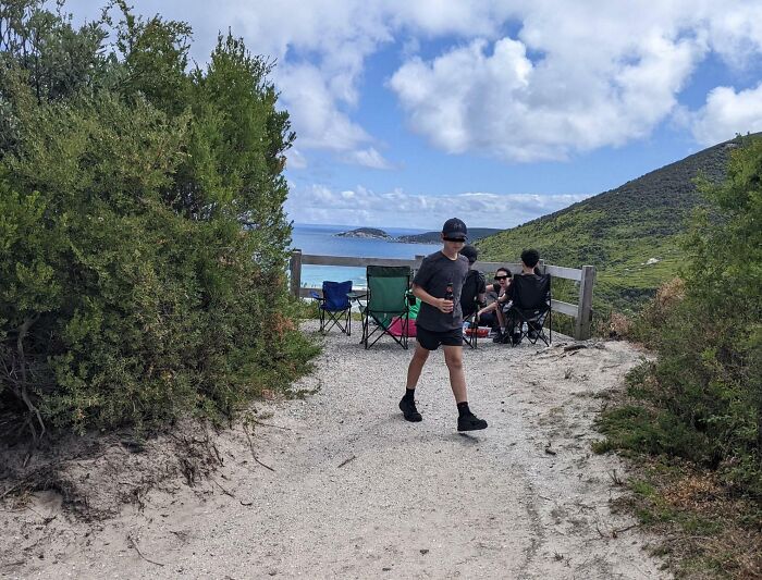 Niño caminando frente a grupo sentado en sillas cerca del mar, mostrando ignorancia sobre normas básicas de educación.