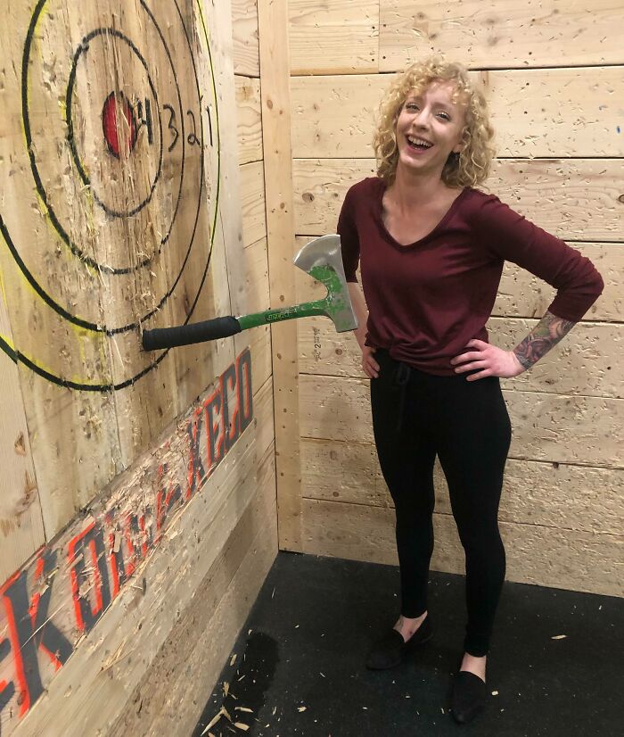 Mujer sonriente junto a un hacha clavada en una diana de madera en actividad de personas con un día peor.