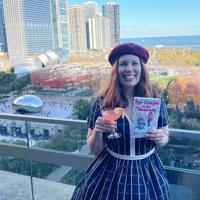 Mujer sonriente con vestido y boina sostiene libro y bebida en balcón con vista urbana, momentos positivos y conmovedores.