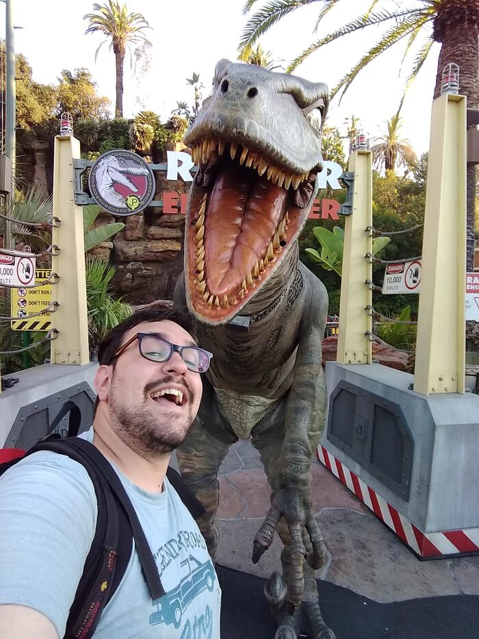 Hombre sonriente junto a un dinosaurio animatrónico en un parque temático, momentos positivos y conmovedores.