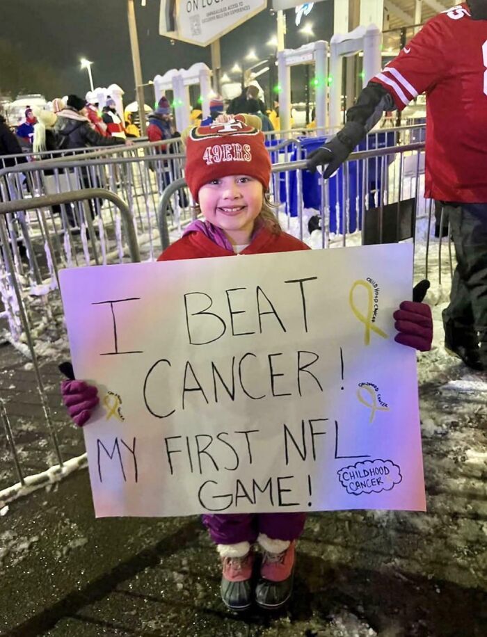 Niña sonriente en partido de NFL con cartel celebrando vencer el cáncer, momento positivo para recobrar la fe en la humanidad.
