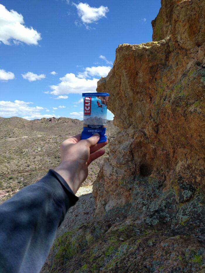 Mano sostiene una barra energética frente a una roca que parece un rostro, foto curiosa y perspectiva original al aire libre.