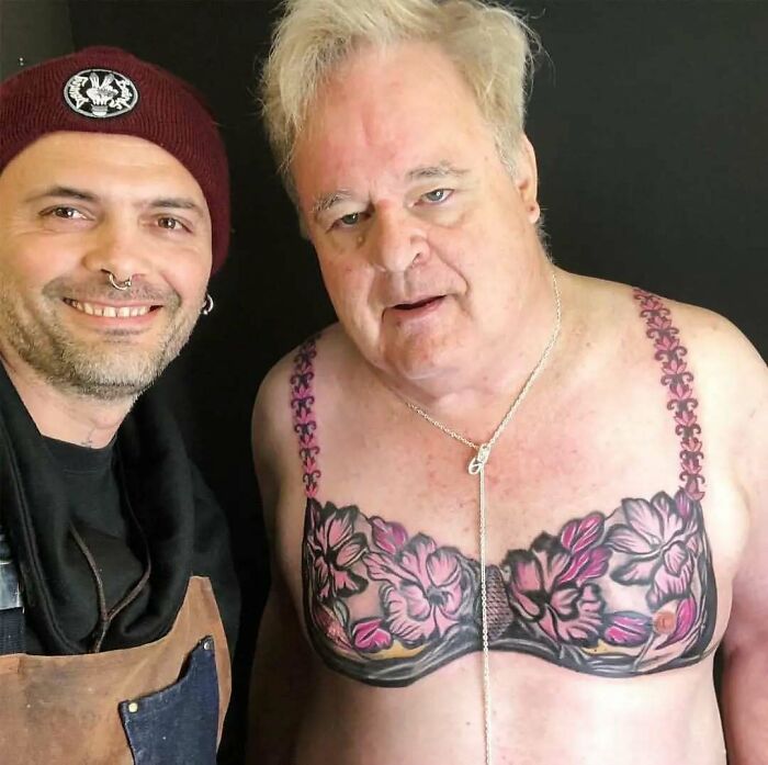 Hombre mayor con tatuaje inusual de flores en el pecho, luciendo sorprendido junto a un joven sonriente.