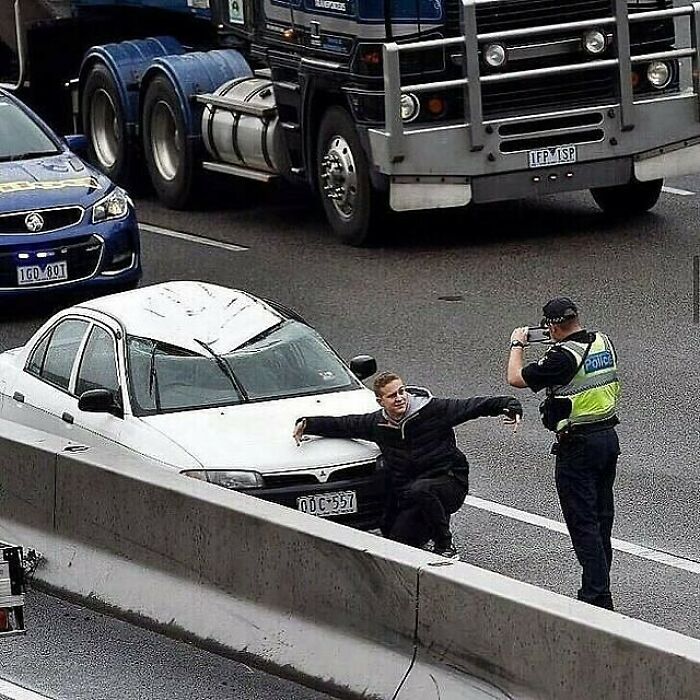 Hombre posando frente a coche blanco en carretera mientras un policía lo fotografía, imagen maldita difícil de explicar.