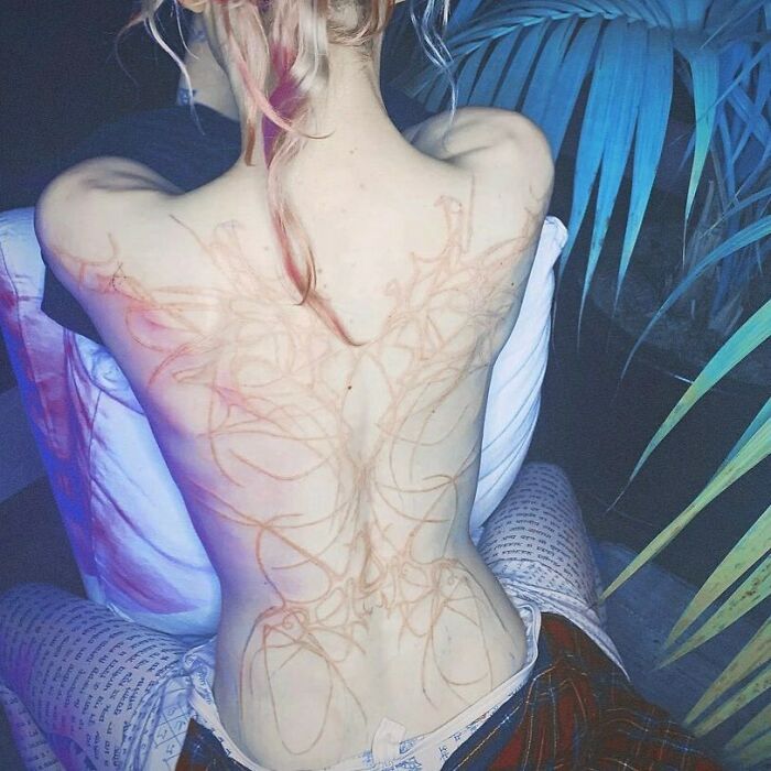 Persona mostrando tatuajes horribles y mal terminados en la espalda bajo luz tenue en un ambiente interior.