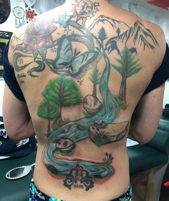 Tatuajes en la espalda con diseño de mariposa, río, árboles y elementos naturales que parecen poco logrados y horribles.