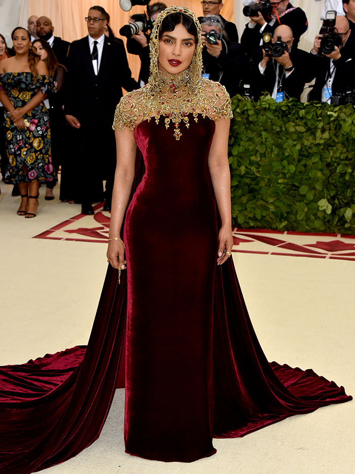 Mujer en vestido burdeos y tocado dorado, rodeada de fotógrafos en la alfombra roja.