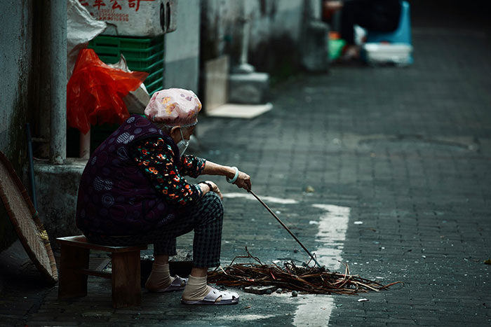 Persona mayor sentada en la calle, interactuando con un montón de ramas, representando un choque cultural.