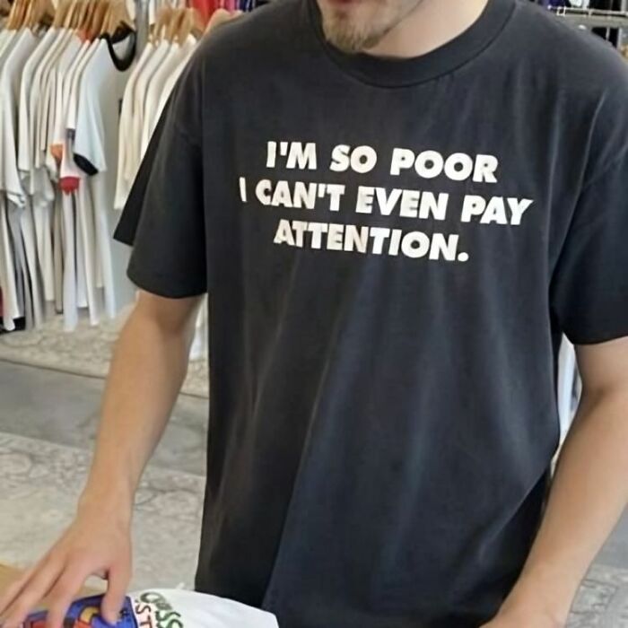 Hombre con camiseta negra divertida con texto en inglés, rodeado de varias camisetas colgadas en tienda de ropa.
