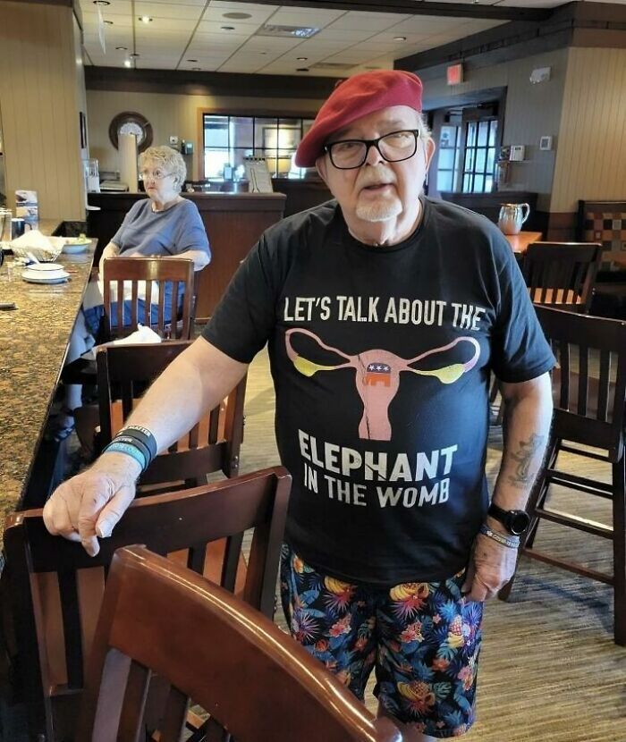 Hombre mayor con camiseta divertida que muestra un diseño ridículo relacionado con un elefante en el útero en un restaurante.