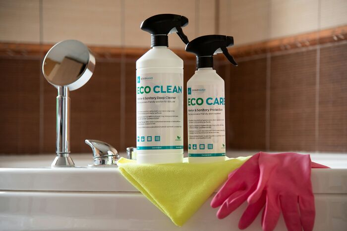 Botellas de productos de limpieza ecológicos con guantes rosas y paño amarillo sobre lavabo en baño.