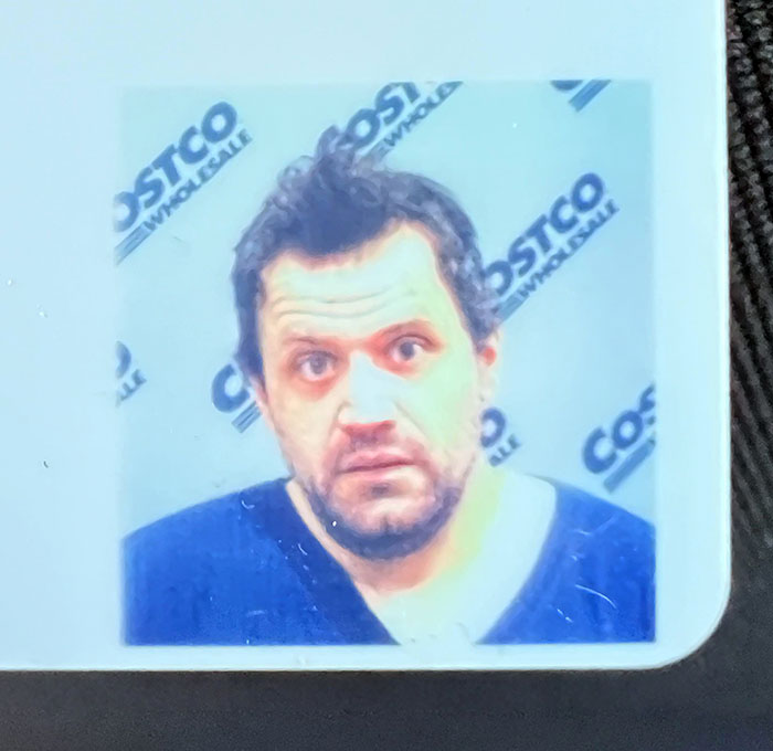 Un novio divertido con camiseta azul en una foto de carné de Costco, mostrando una expresión graciosa.