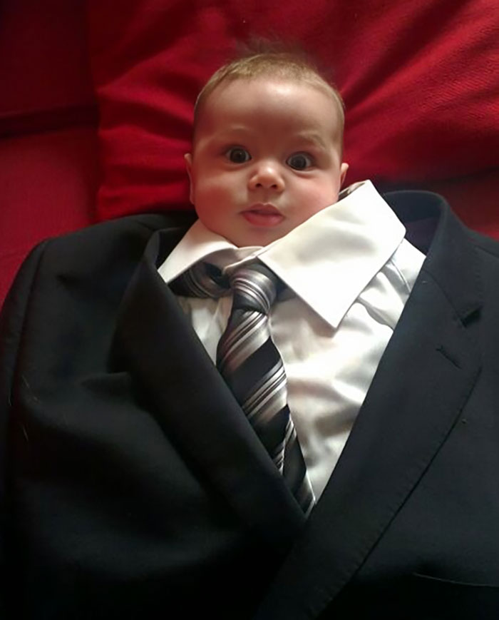 Bebé con expresión divertida, vestido con traje adulto y corbata, sobre fondo rojo.