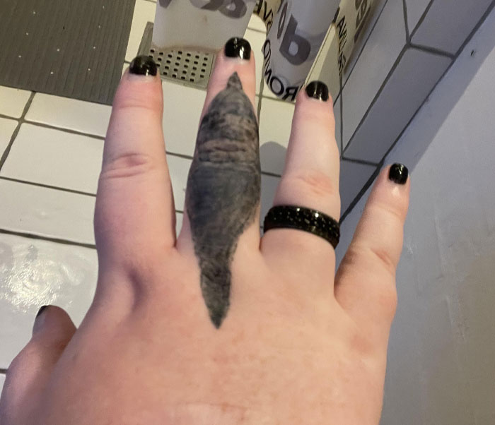 Mano con esmalte negro y un tatuaje mal hecho en el dedo medio, ejemplo de tatuajes que fueron una mala elección.