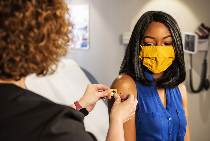 Persona con mascarilla amarilla recibiendo una vacuna de una profesional de salud en un consultorio médico, datos que te harán cuestionar.