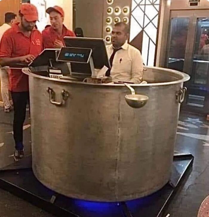 Hombre sentado dentro de olla gigante con computadora mientras otros dos hombres trabajan cerca en restaurante, imagen maldita divertida.