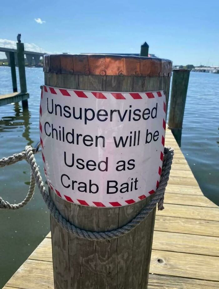 Señal humorística en muelle que advierte sobre niños sin supervisión usados como cebo para cangrejos, imágenes malditas y bendecidas.