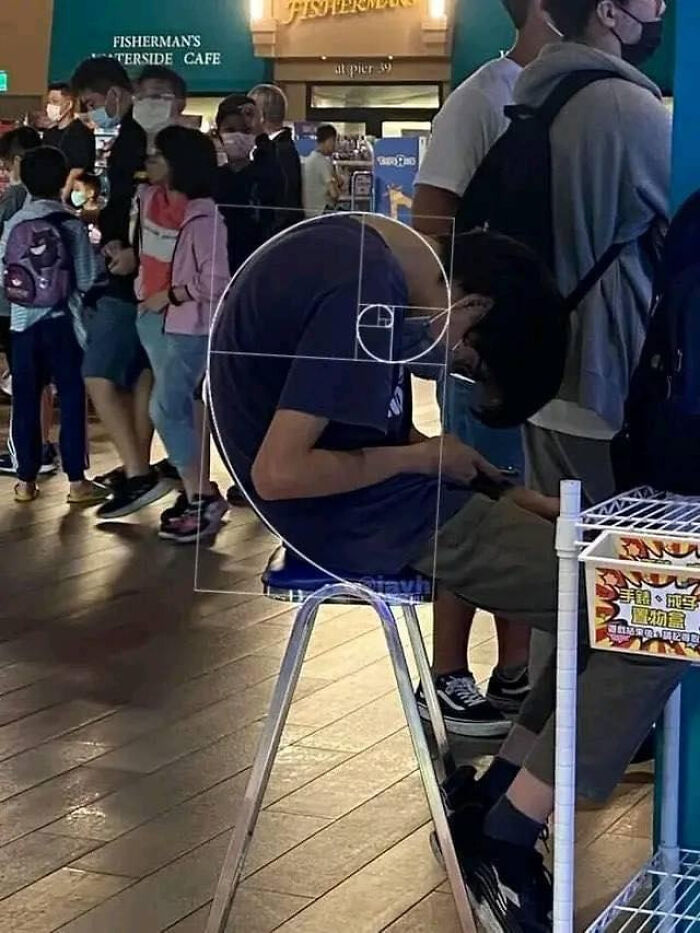 Persona sentada encorvada en taburete con postura que forma espiral, imagen bendecida y maldita en espacio público concurrido.