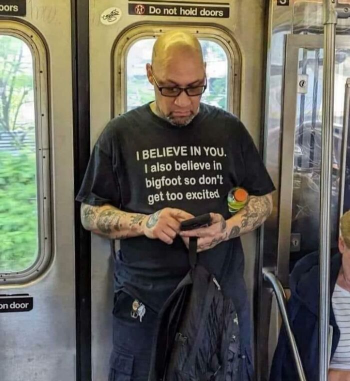 Hombre con tatuajes en camiseta con texto curioso, usando celular en transporte público, imagen bendecida y maldita al mismo tiempo.
