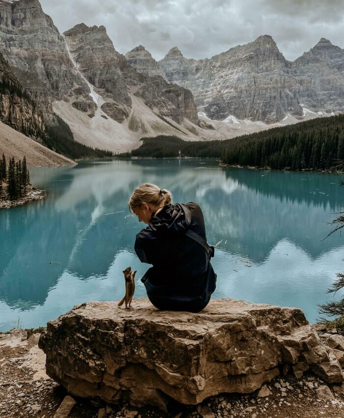 Mujer sentada en roca junto a lago y montañas, interactuando con ardilla cerca de coincidencias increíbles en la naturaleza.