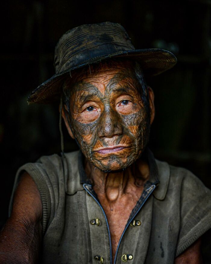 Retrato elocuente de persona mayor con tatuajes faciales tradicionales, usando sombrero y camisa en ambiente oscuro.