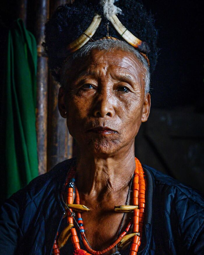Retrato elocuente de persona mayor con adornos tradicionales y expresión seria en luz tenue, mostrando detalles de su piel y cultura.
