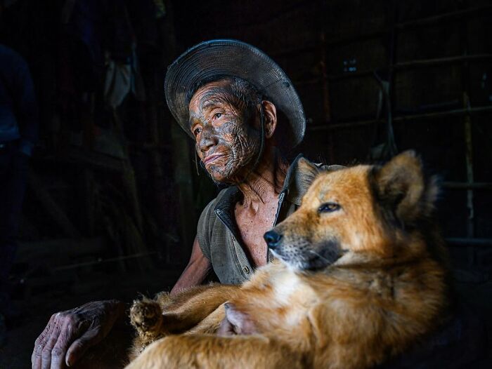 Hombre mayor con tatuajes faciales tradicionales y sombrero, acompañado de un perro en el interior oscuro, retrato elocuente.
