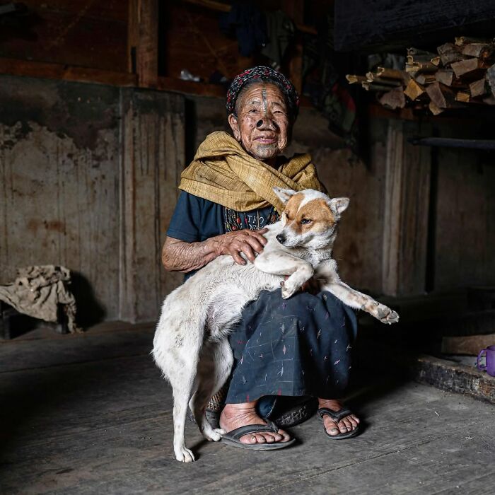 Retrato elocuente de persona mayor con perra en ambiente rústico, mostrando la conexión y vida cotidiana alrededor del mundo.