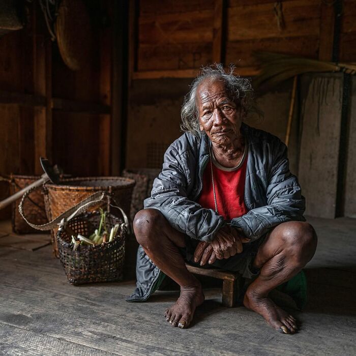 Retrato elocuente de persona mayor sentada en suelo de madera, con cestas y herramientas en entorno rústico.