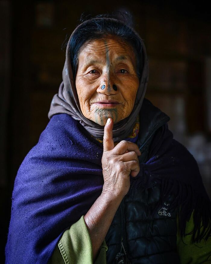 Retrato elocuente de mujer mayor con tatuajes faciales tradicionales en su rostro, vestida con chal azul y bufanda.