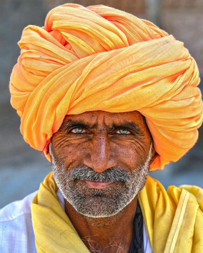 Retrato elocuente de hombre mayor con turbante naranja y ojos claros, mostrando arrugas y expresión serena.