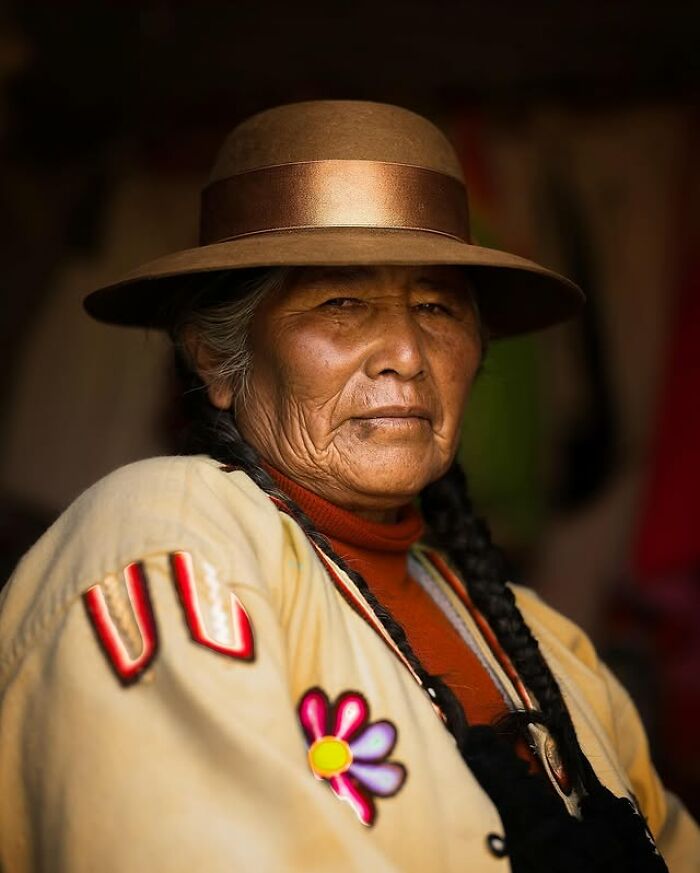 Retrato elocuente de persona mayor con sombrero marrón, mostrando arrugas y expresión serena en ambiente oscuro.