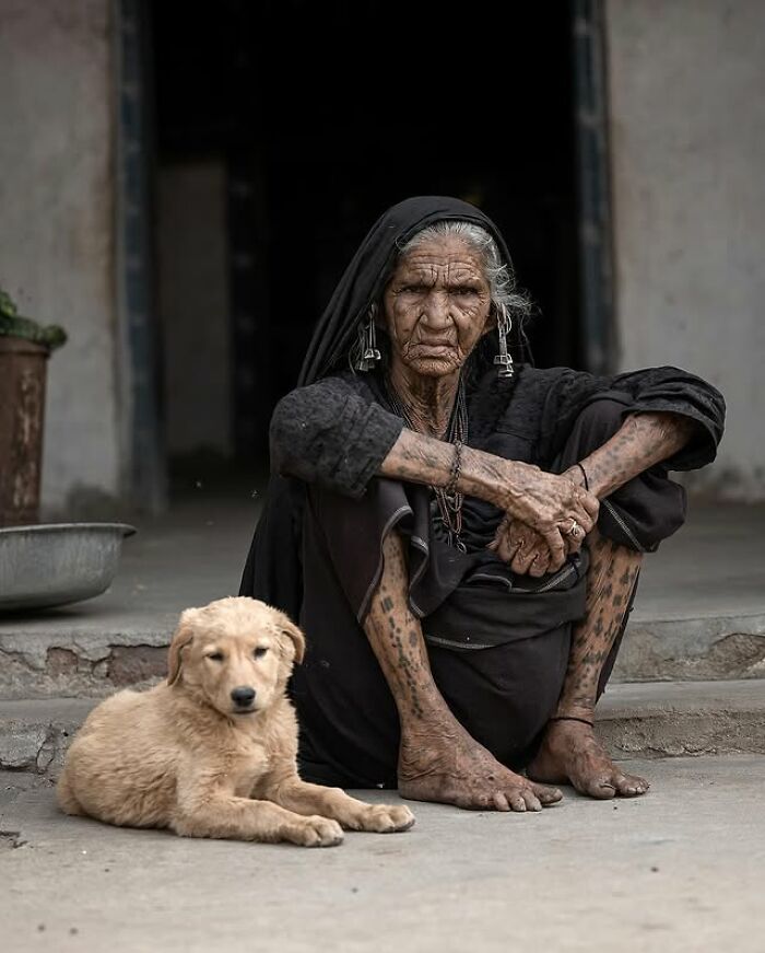 Retrato elocuente de persona mayor con tatuajes tradicionales sentada junto a un cachorro en un entorno rural.